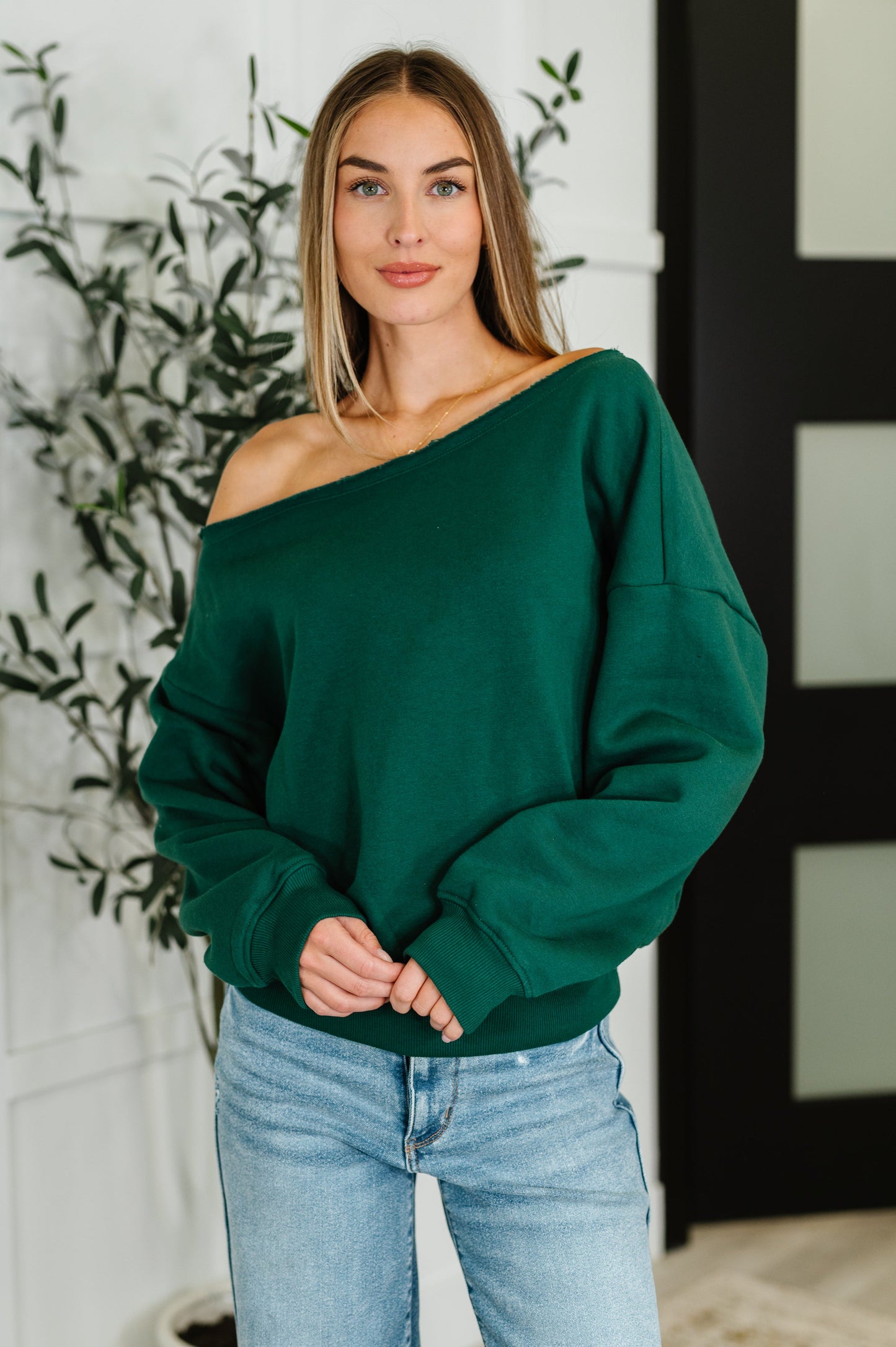 Woman wearing a green off-shoulder long sleeve shirt top and light blue jeans indoors.