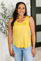 Woman wearing a yellow tank top and blue jeans standing indoors with a plant in the background