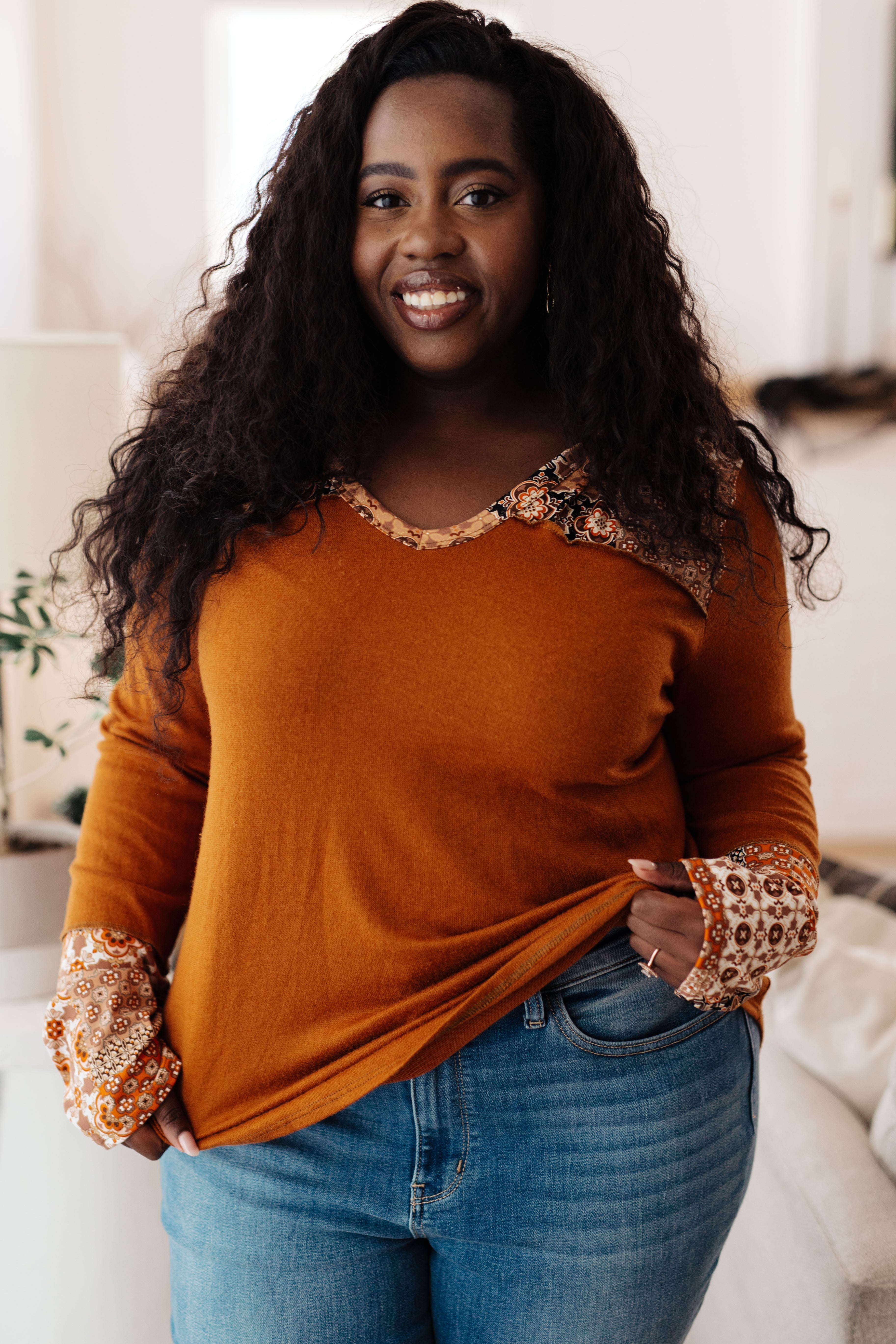 Woman wearing a rust-colored long-sleeve top with patterned cuffs and hem, standing in a bright room.
