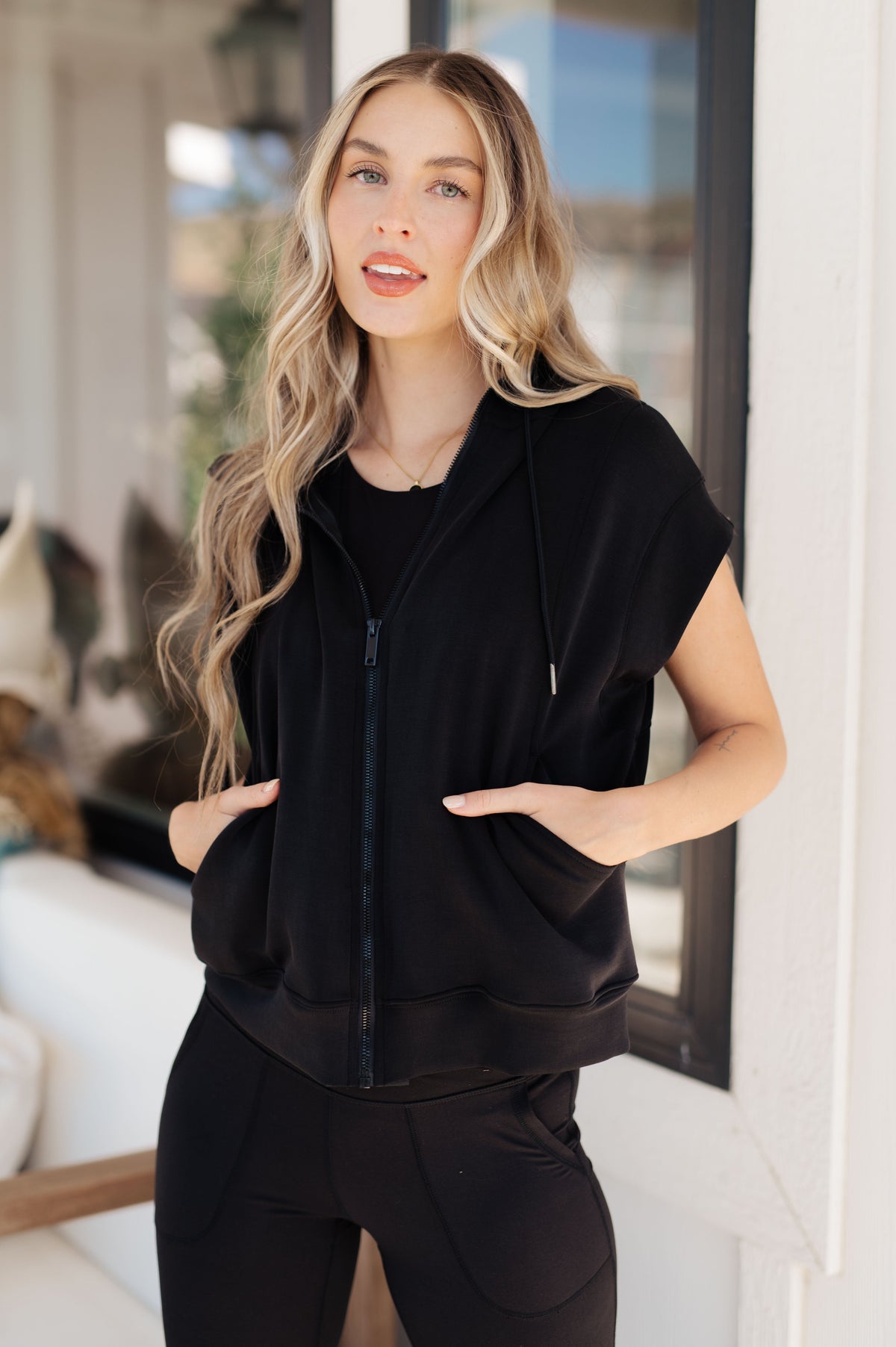 Model wearing a black sleeveless hoodie with a zip front and pockets.
