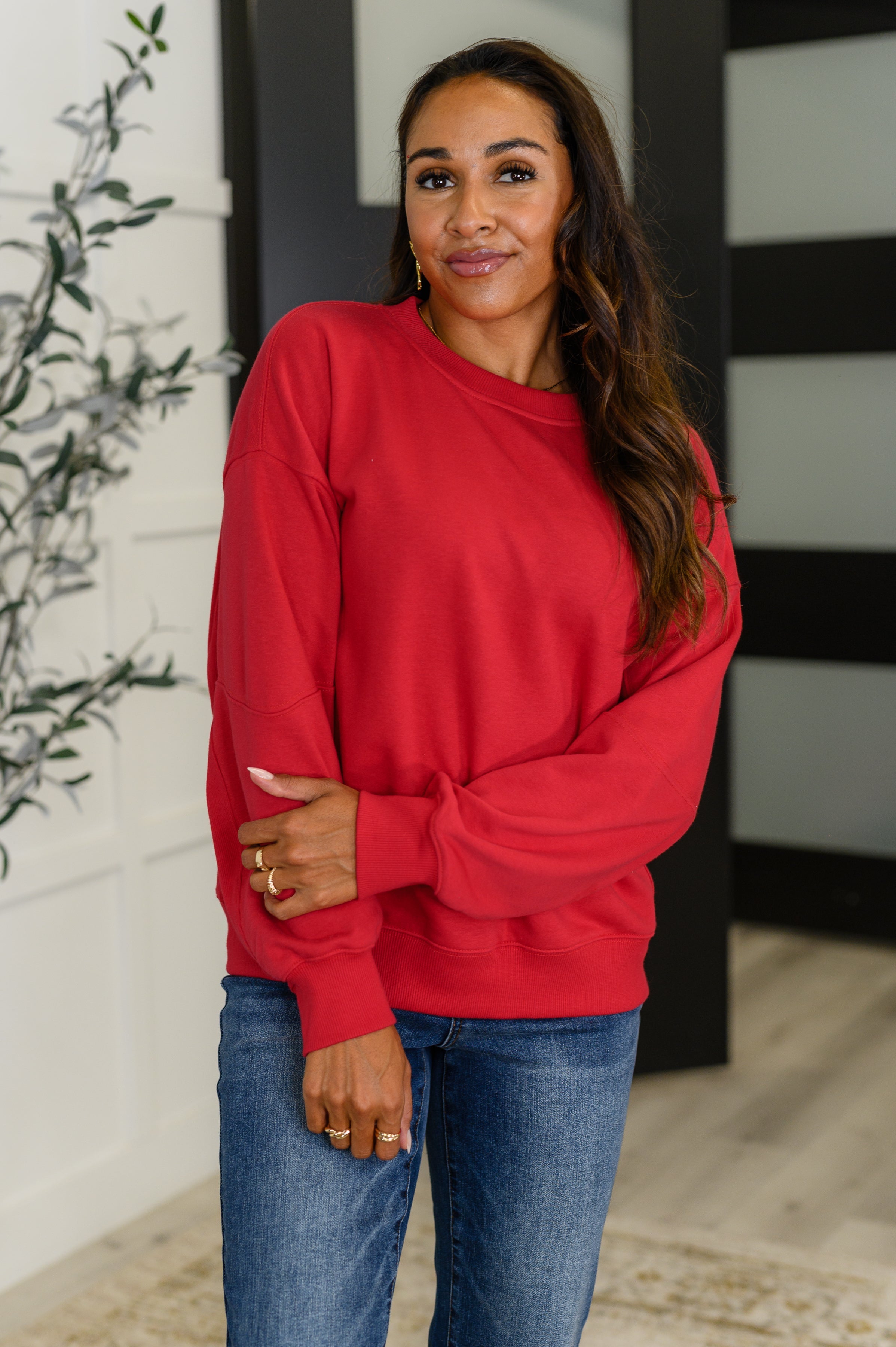 Woman wearing a red sweatshirt top and blue jeans standing indoors.