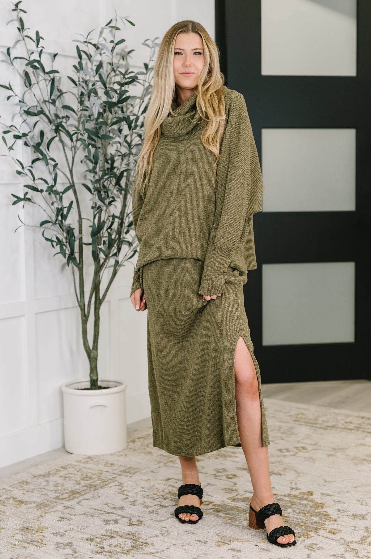 A woman wearing an olive waffle knit set with a cowl neck and side slits, standing in a room.