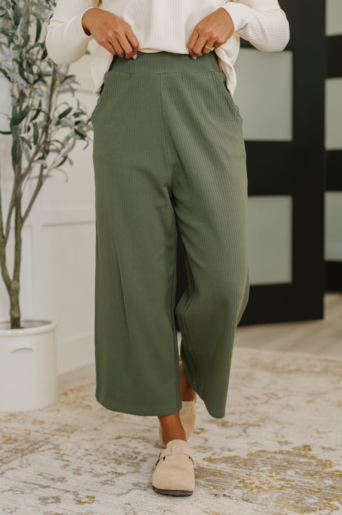 Woman wearing green pants and beige shoes in a room with a plant and decorative wall.