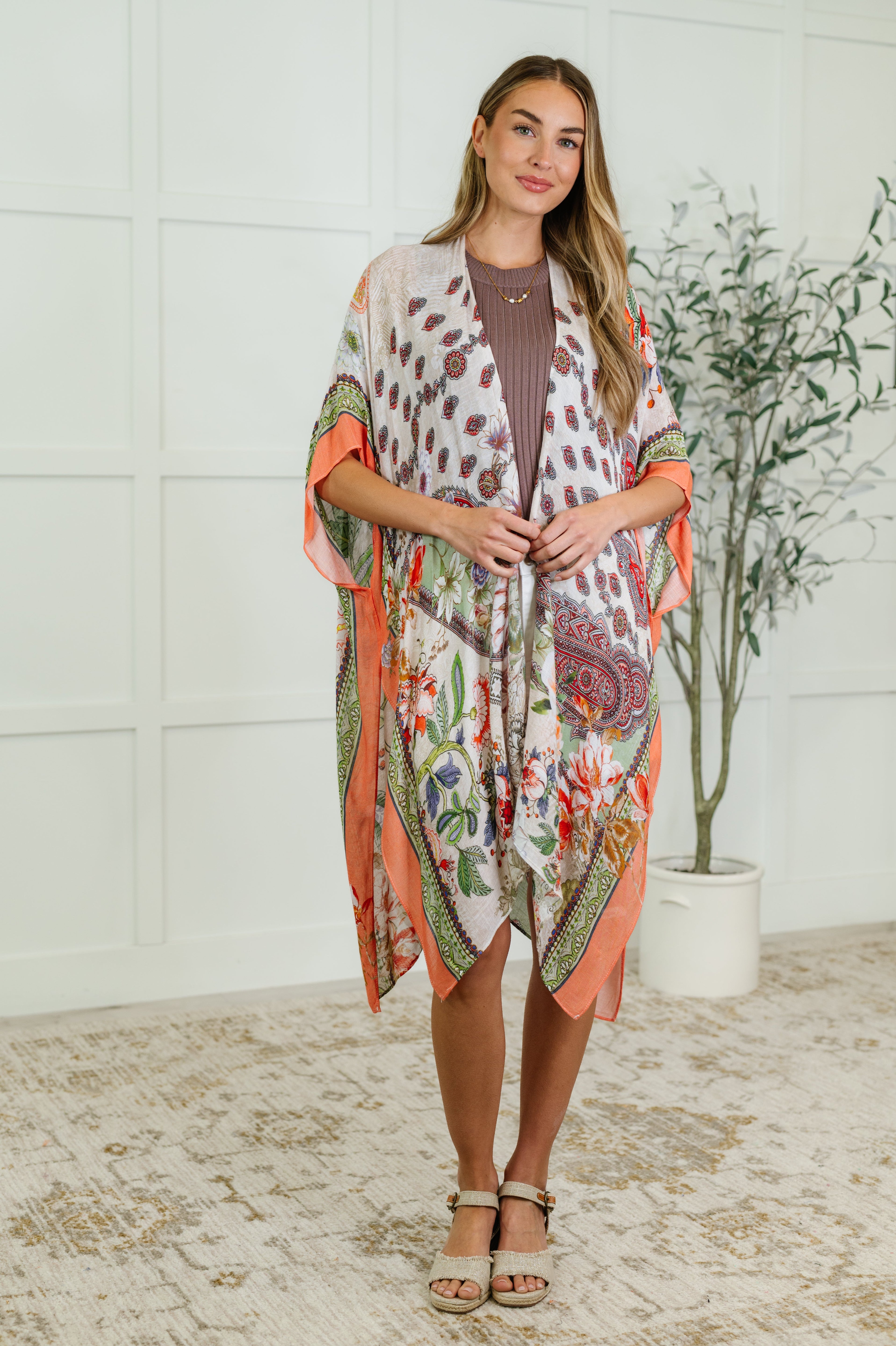 Woman wearing a multicolored open front kimono with floral print, paired with a brown top and beige sandals.