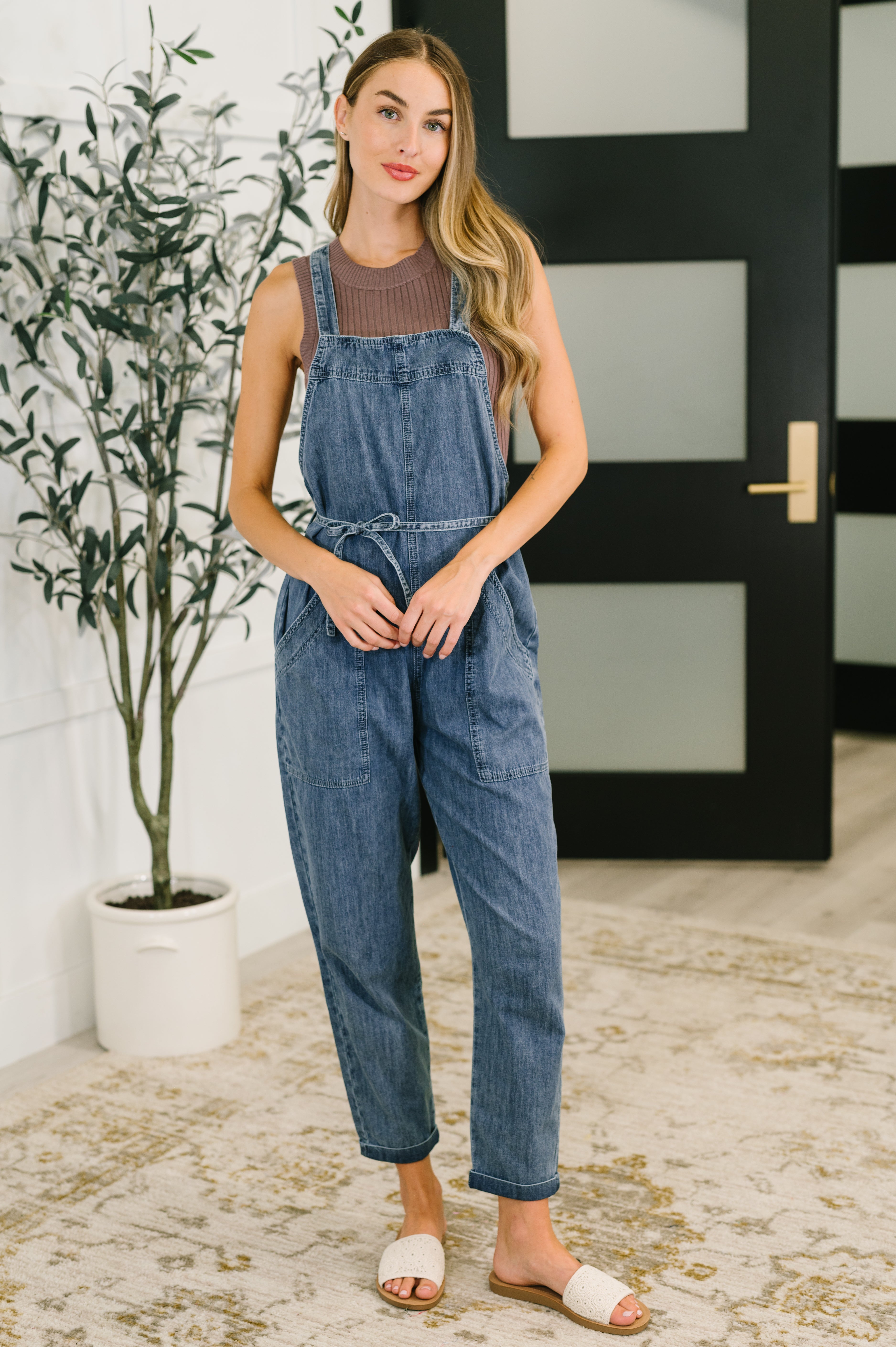 Woman wearing a denim jumpsuit standing indoors with a plant and door in the background