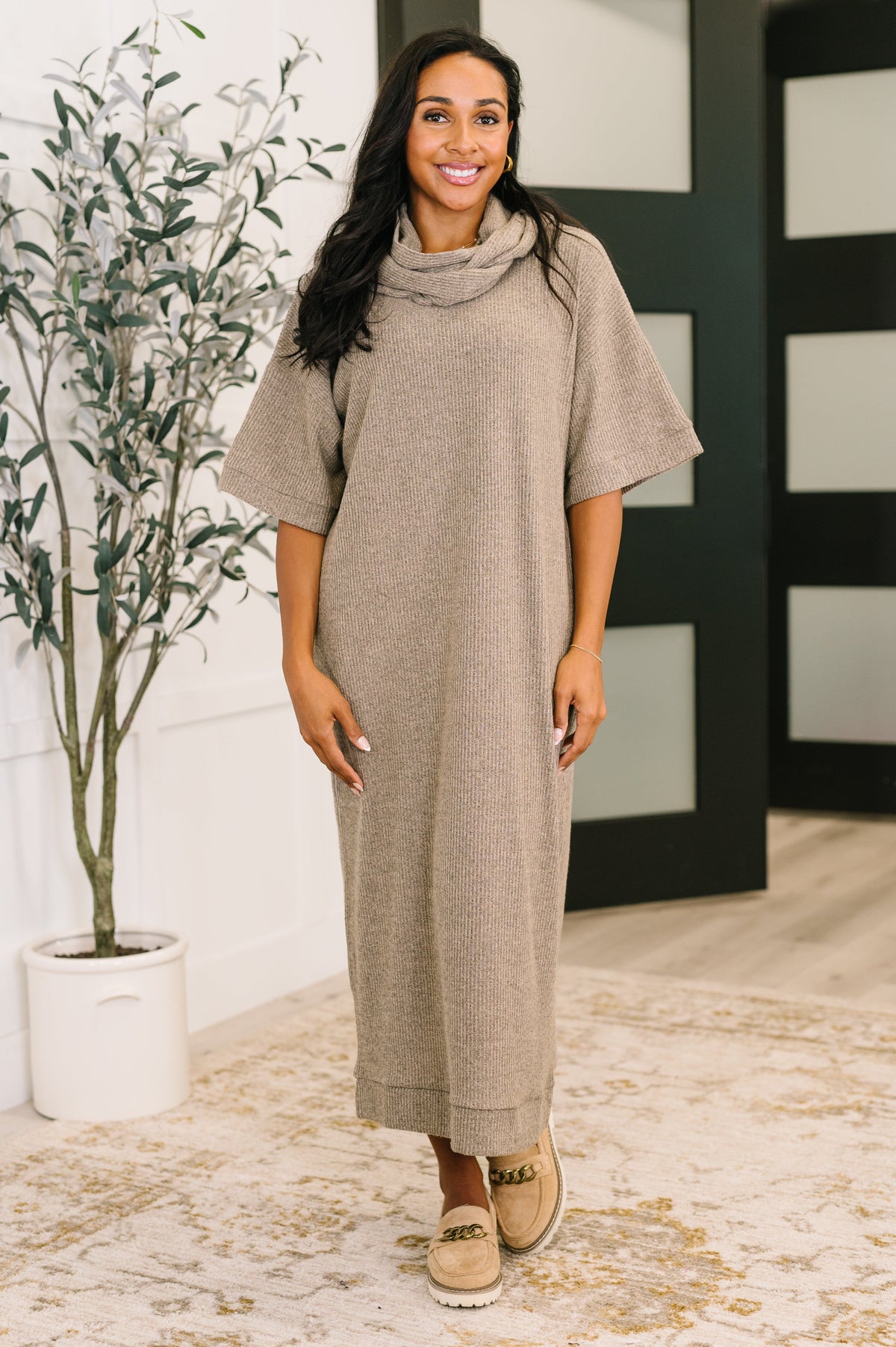 A woman wearing a light coco ribbed knit dress with a V-neckline and attached neck tie, featuring drop shoulders and side pockets.