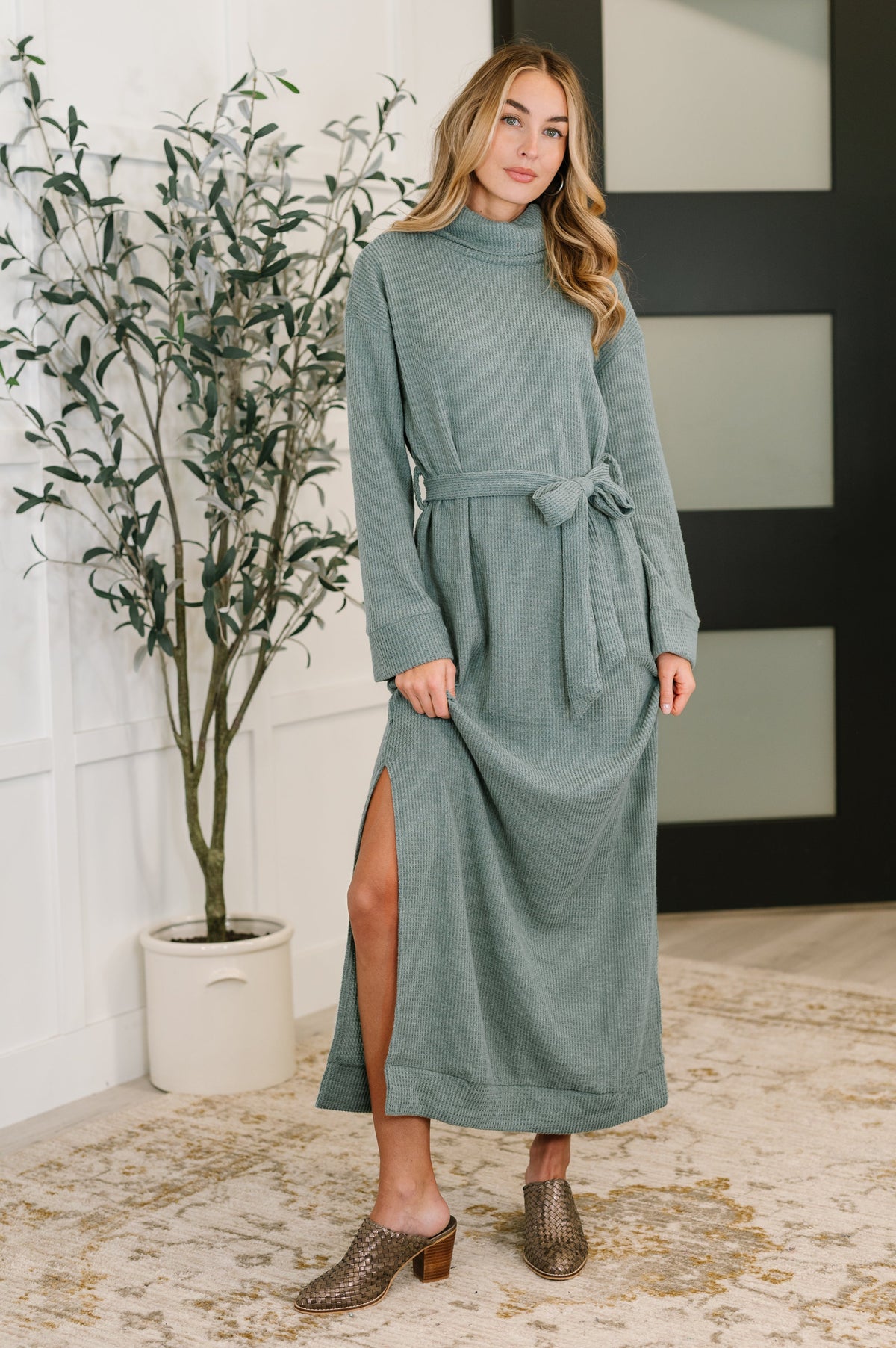 A model wearing a soft teal knit dress with a turtleneck, long sleeves, waist tie, and side slits.