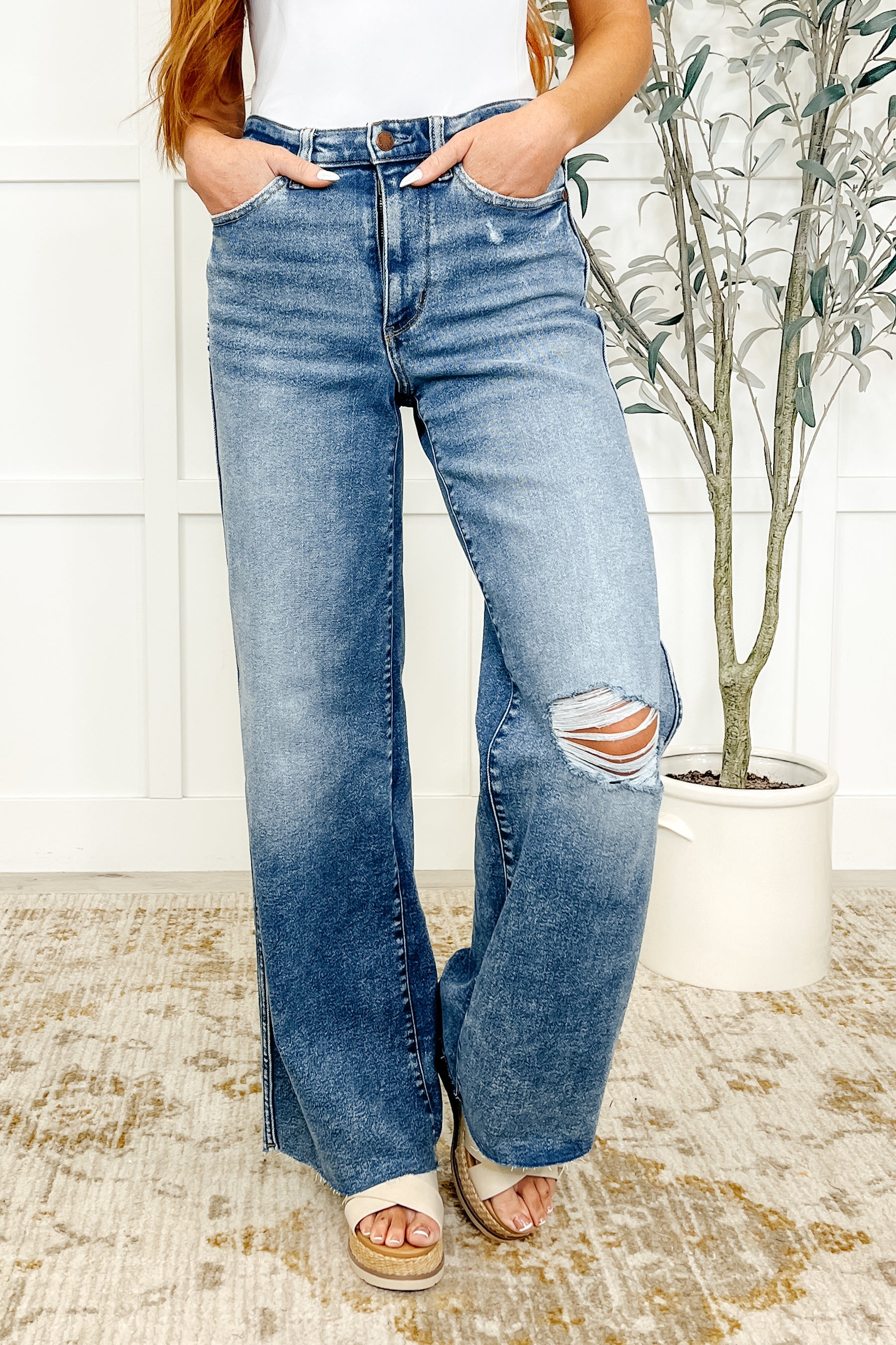 Woman wearing a pair of high-waisted, vintage wash wide leg jeans with distressed knees and a raw hem. The jeans are paired with a white top.