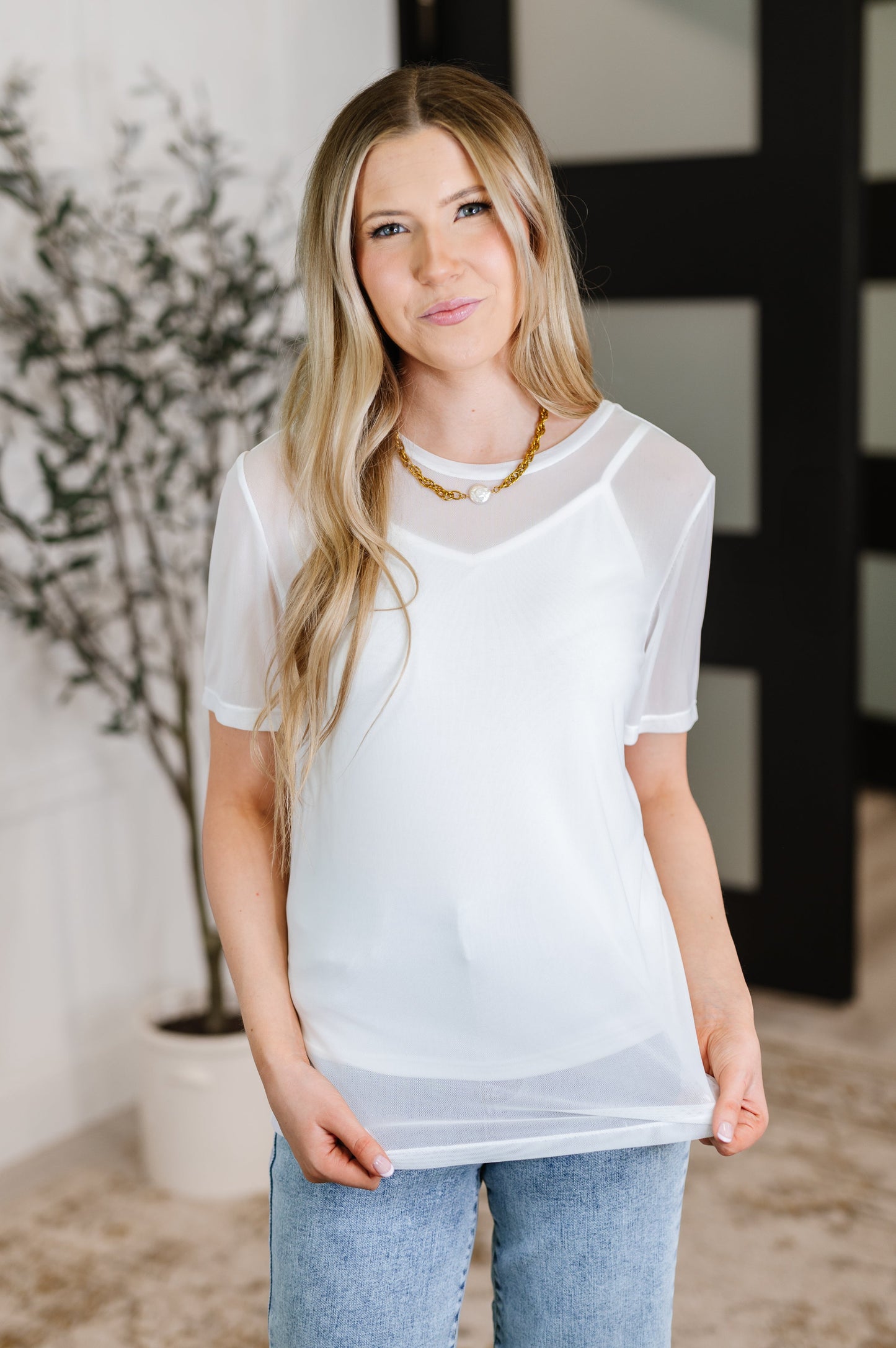 Woman wearing a sheer white top and jeans indoors with a plant in the background