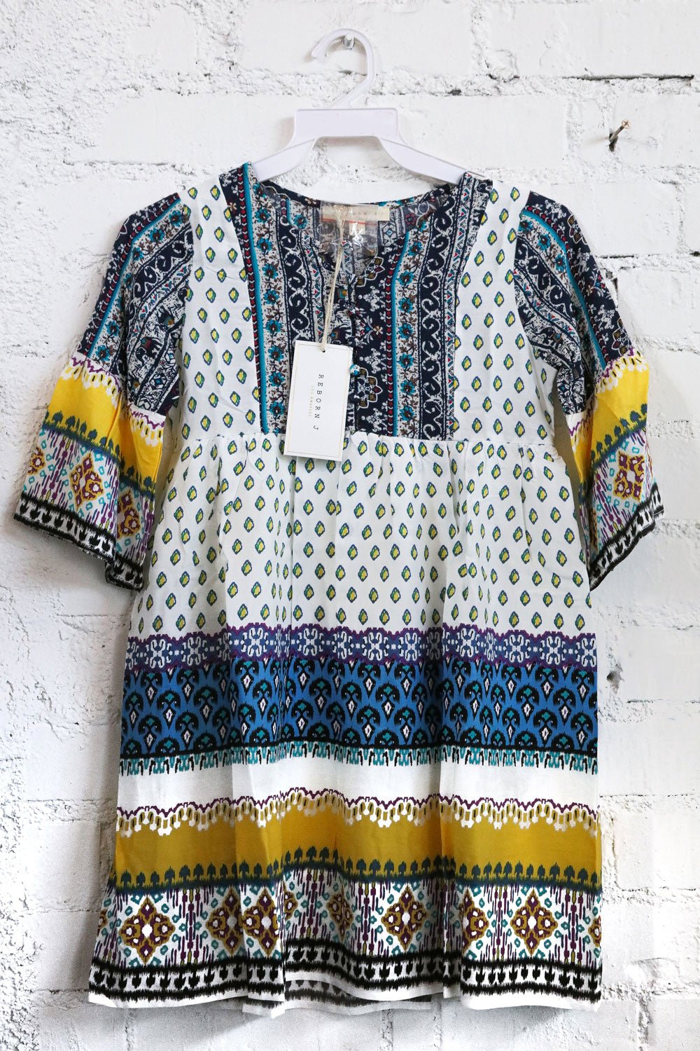 Kids colorful patterned dress on a hanger against a white brick wall.