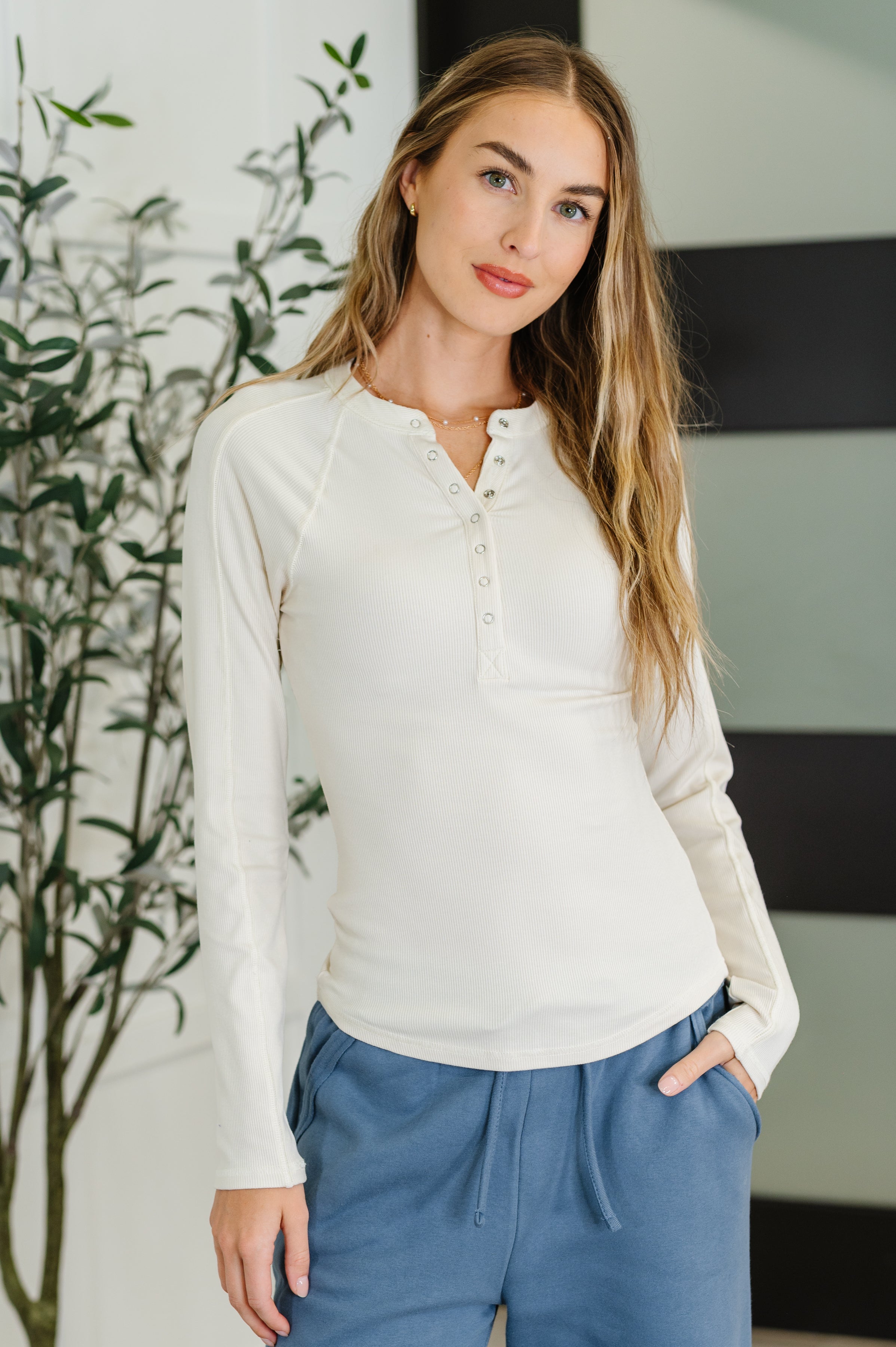 Woman wearing a white long-sleeve shirt and blue pants standing indoors with a plant in the background