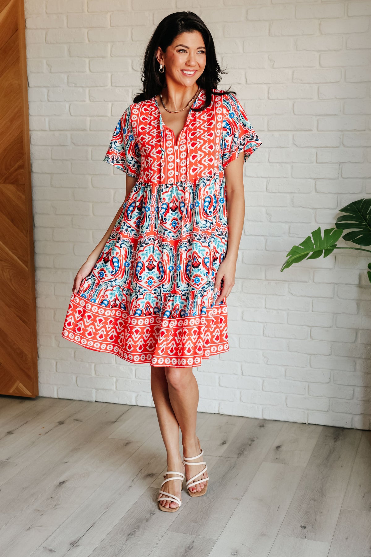 A woman standing in a room wearing a red and blue floral print dress with a v-neckline, flutter sleeves, and a tiered skirt. She has accessorized with neck ties and faux button accents.