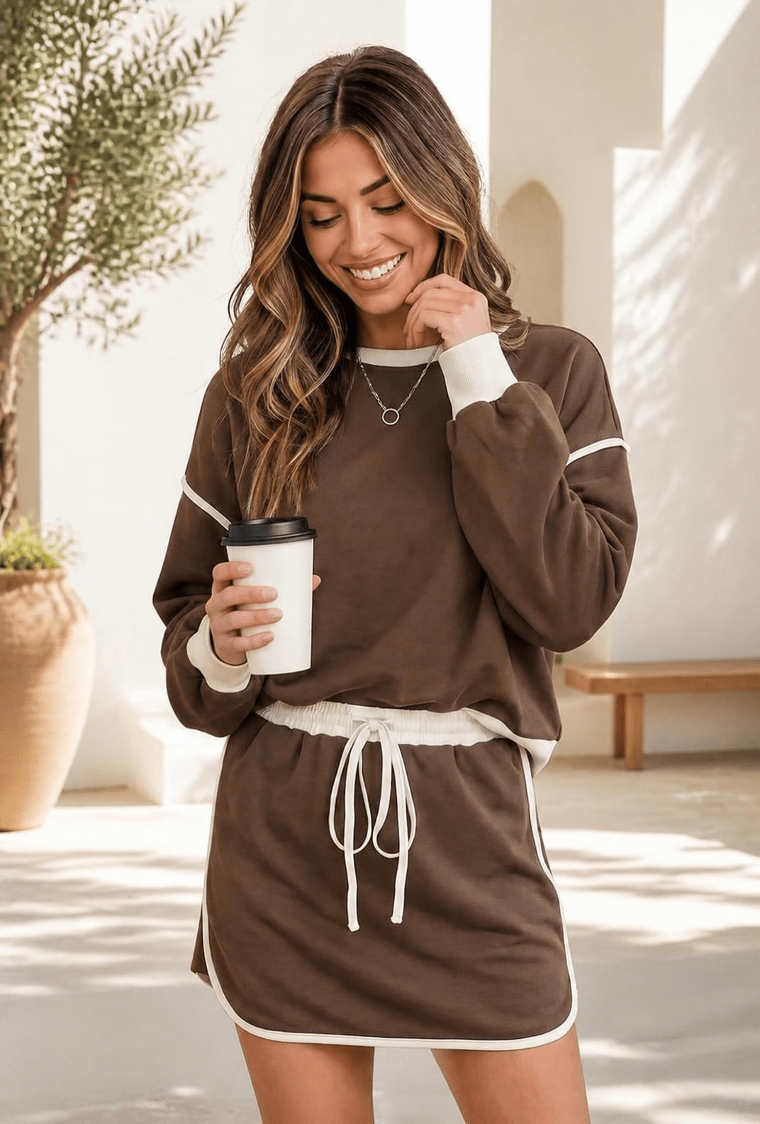 A woman wearing a mocha brown sweatshirt and mini skort set with cream contrast trim, holding a coffee cup.