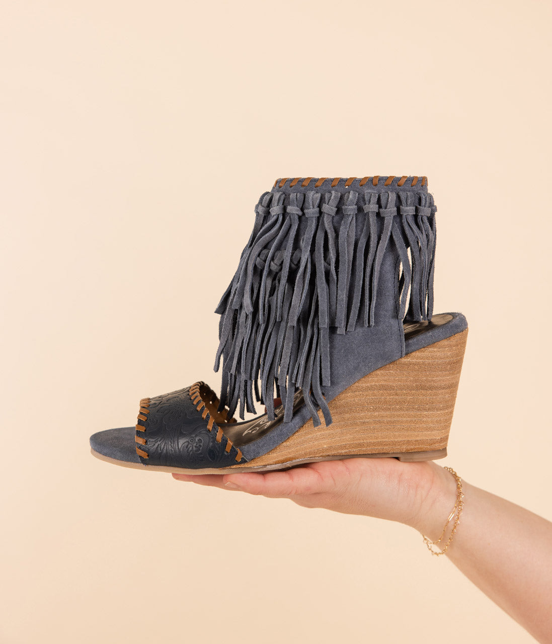 A denim blue wedge sandal with fringe ankle detail held in a hand against a beige background.