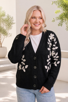 A woman wearing a black cardigan with cream floral embroidery, buttoned in front, with ribbed cuffs and a relaxed fit.