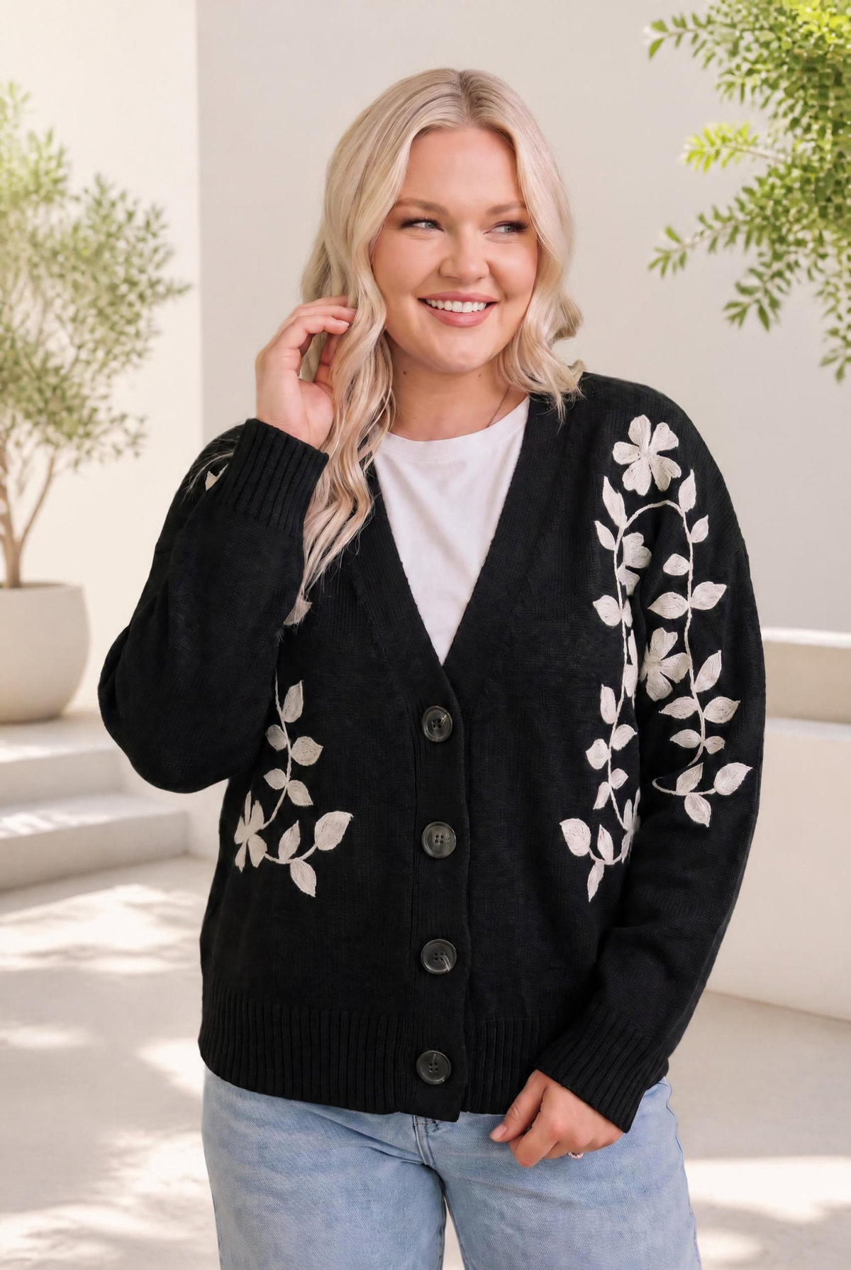 A woman wearing a black cardigan with cream floral embroidery, buttoned in front, with ribbed cuffs and a relaxed fit.