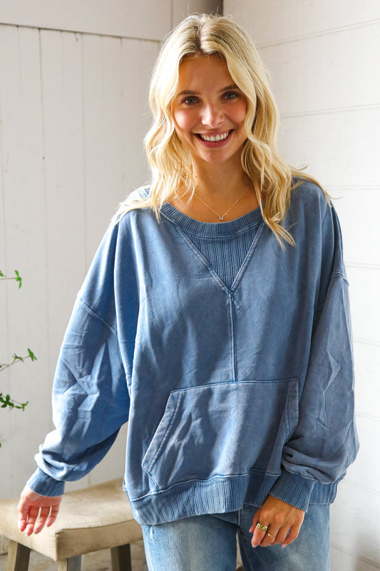 Woman wearing a blue sweatshirt and jeans standing against a white wall.