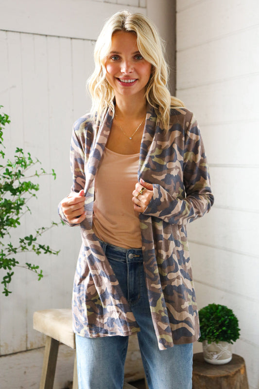 Woman wearing a plaid cardigan shirt over a beige top and jeans, standing indoors with plants in the background.