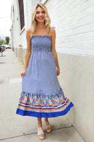 Woman wearing a blue floral dress with colorful trim standing against a white wall.