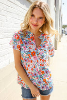 Woman wearing a colorful floral blouse standing against a white brick wall.