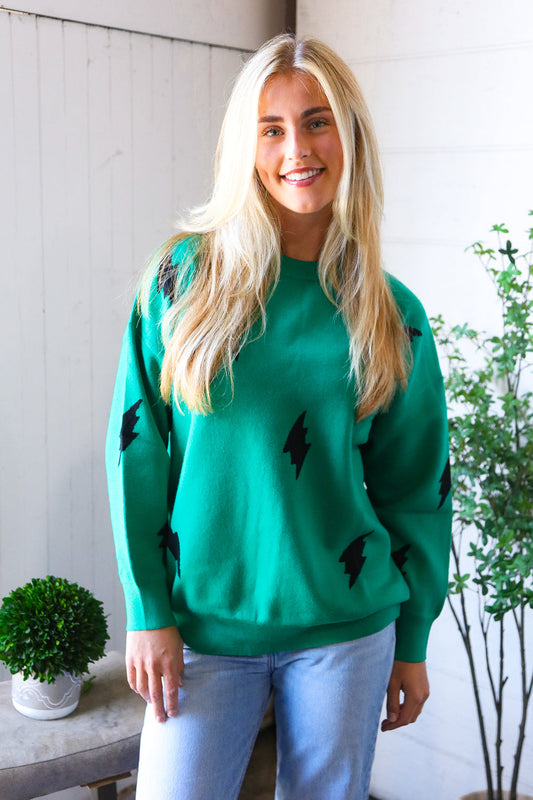 Woman wearing a green long sleeve shirt top with black designs, standing indoors with plants in the background.