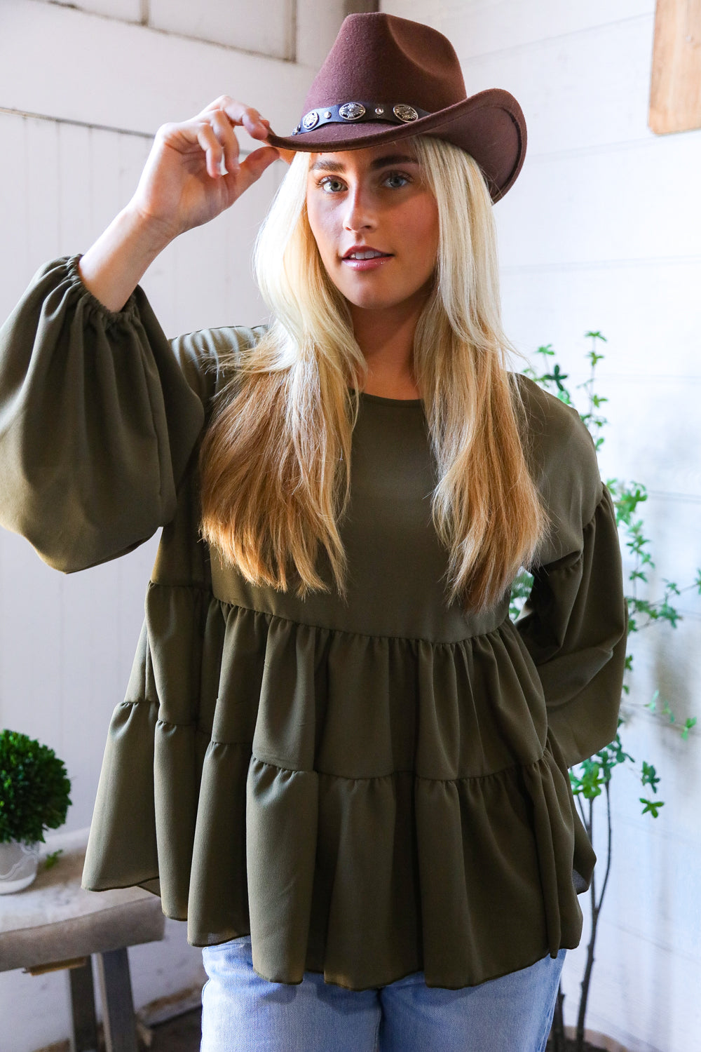 Woman wearing an olive green tiered blouse and brown hat indoors.