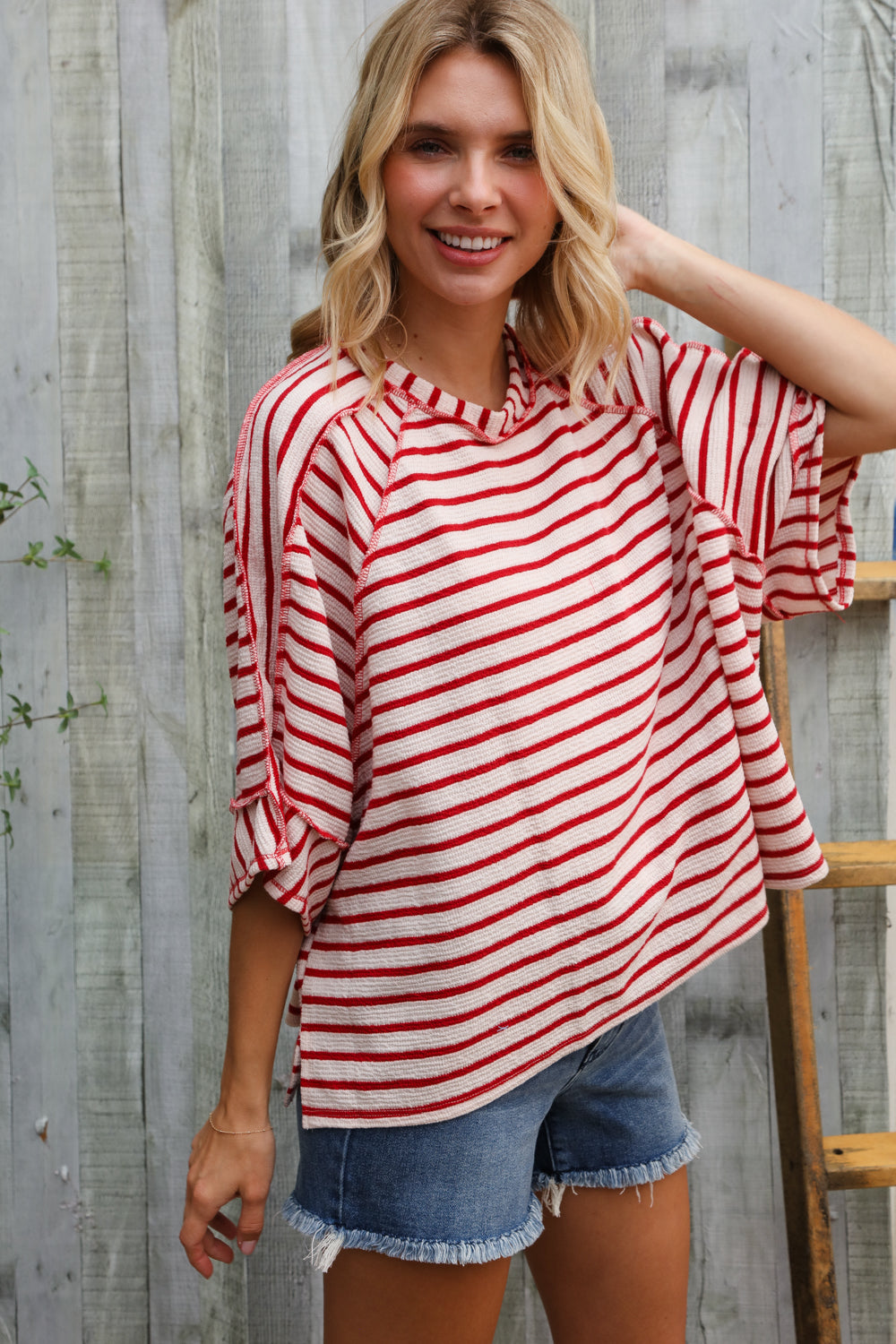 Woman wearing a red and white striped shirt and denim shorts standing against a wooden fence.