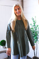 Woman wearing a long green tunic shirt withs stripes s standing indoors with plants in the background