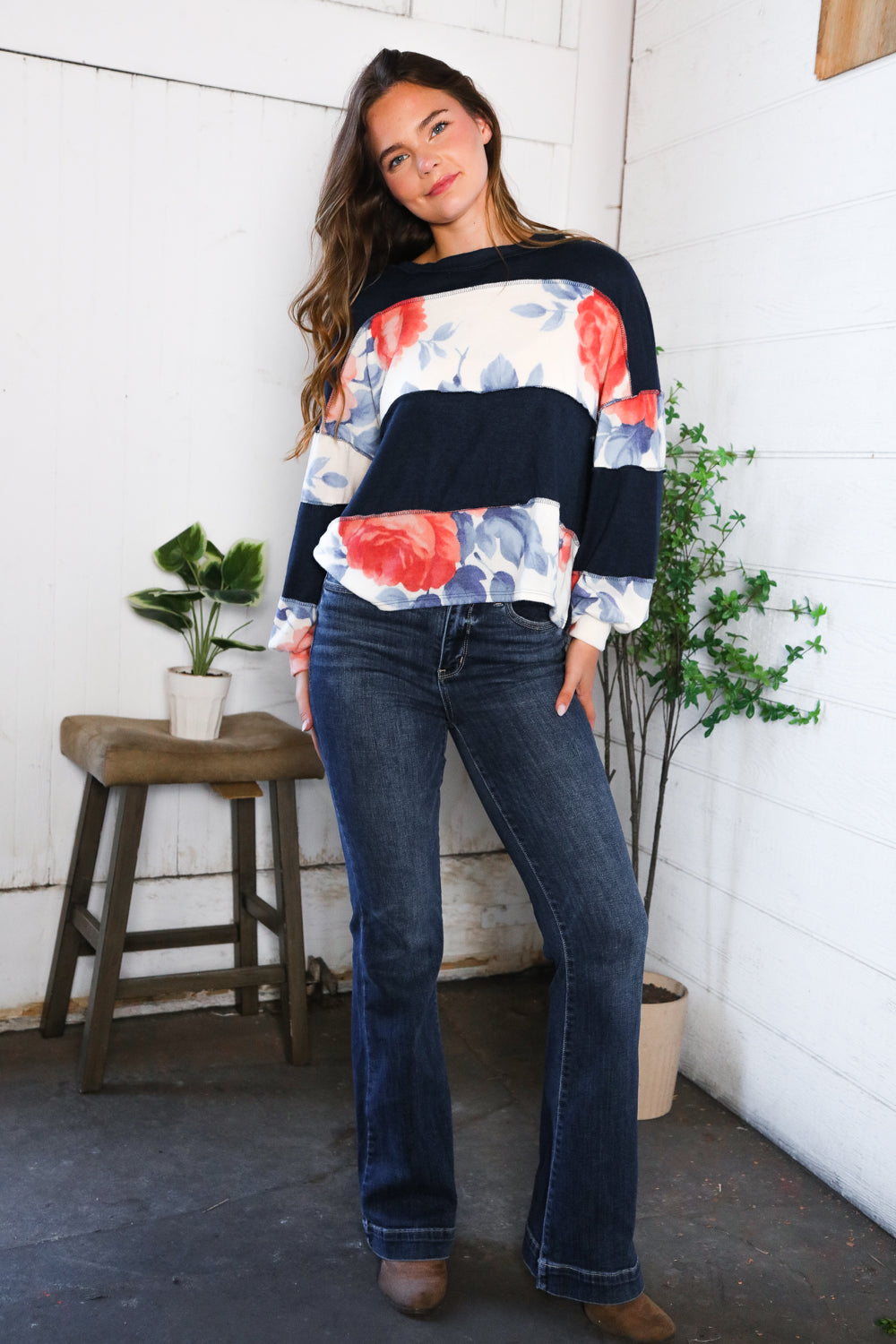 Woman wearing a floral long sleeves shirt and jeans standing in front of a white wall with plants.