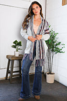 Woman wearing a patterned cardigan and jeans standing indoors with plants and a stool in the background.