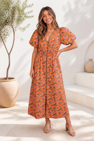 A woman wearing a floral dress with puff sleeves and a smocked waist in orange and lavender print on an olive base.