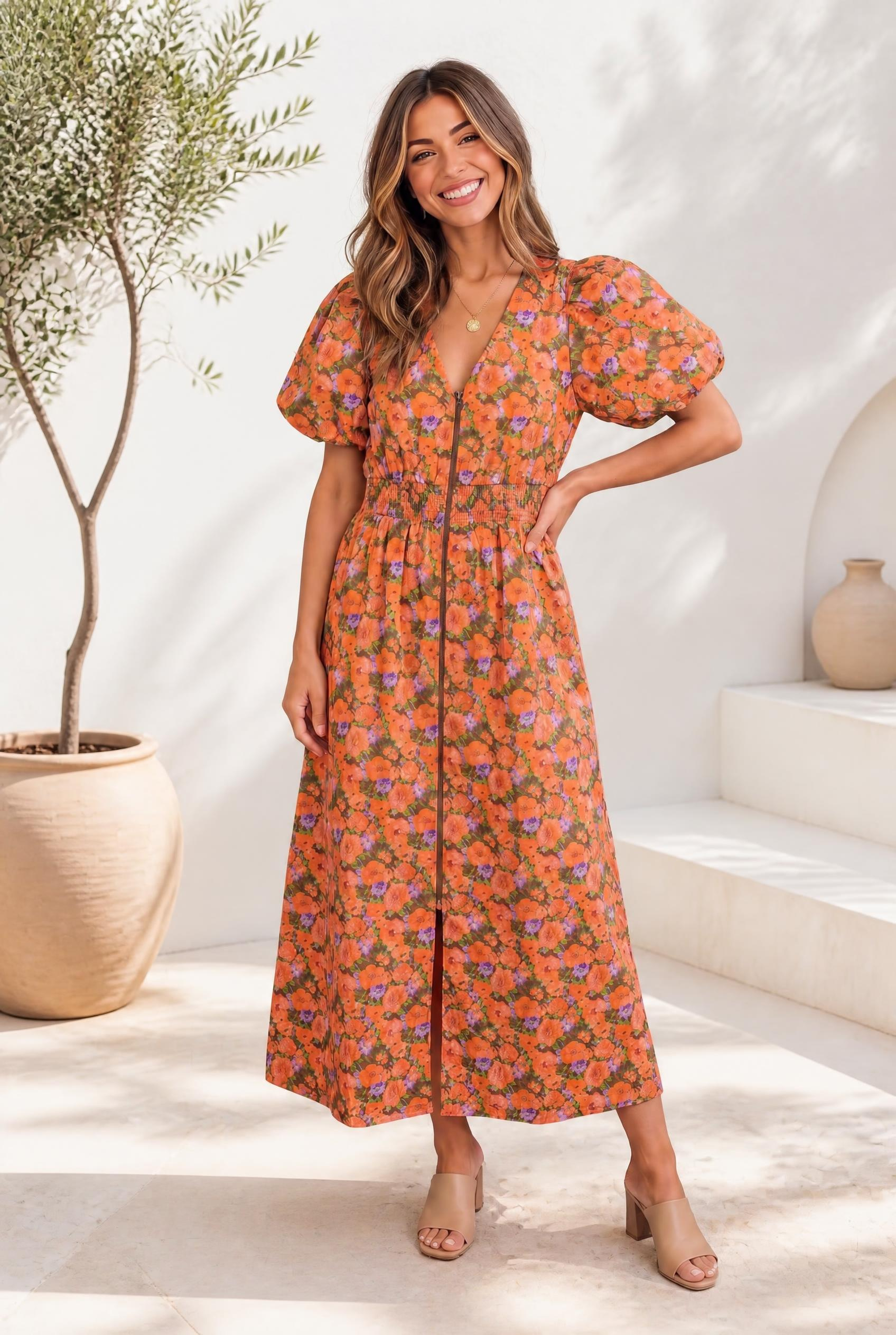 A woman wearing a floral dress with puff sleeves and a smocked waist in orange and lavender print on an olive base.