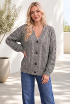 A woman wearing a chunky olive cable knit cardigan with dark buttons and a V-neckline, paired with jeans.