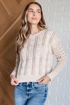 Woman wearing a beige knitted sweater and blue jeans against a white brick wall.