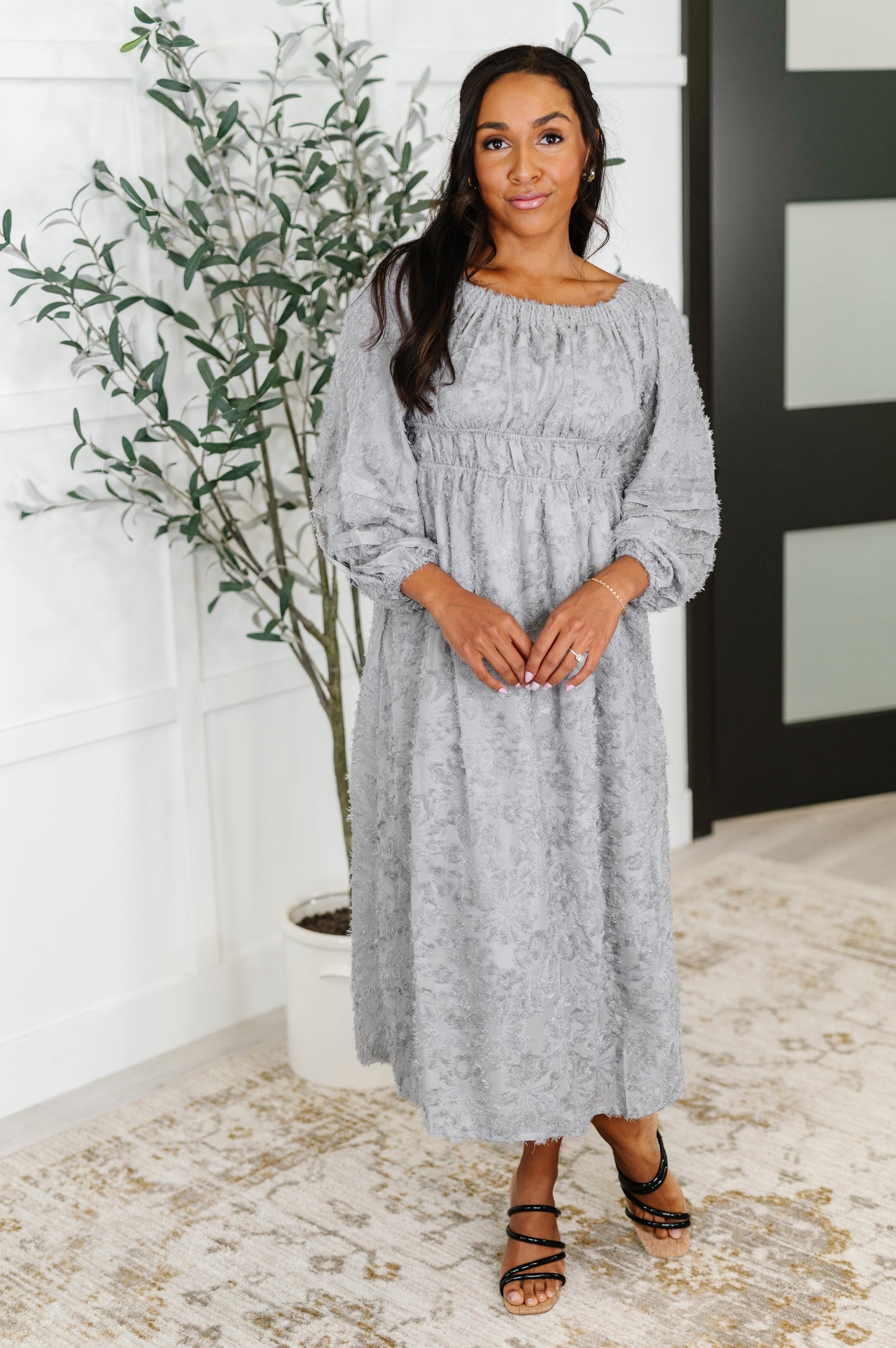 Woman wearing a gray dress standing indoors with a plant in the background