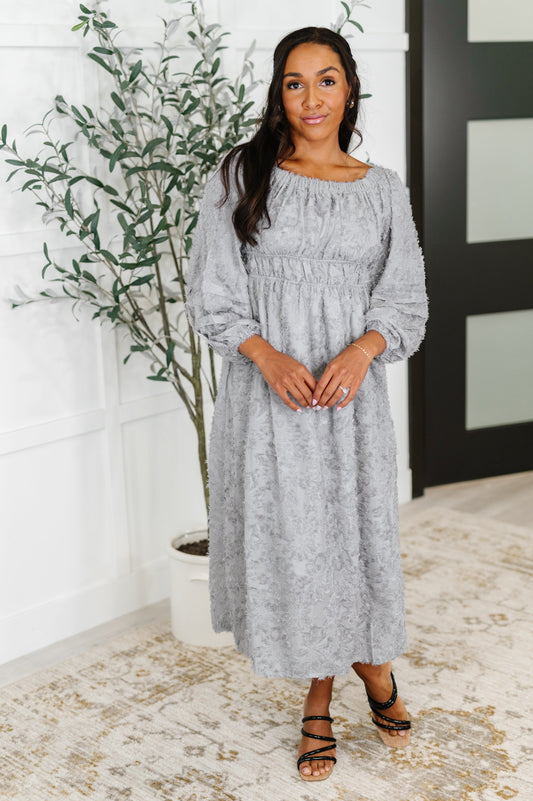 Woman wearing a gray dress standing indoors with a plant in the background
