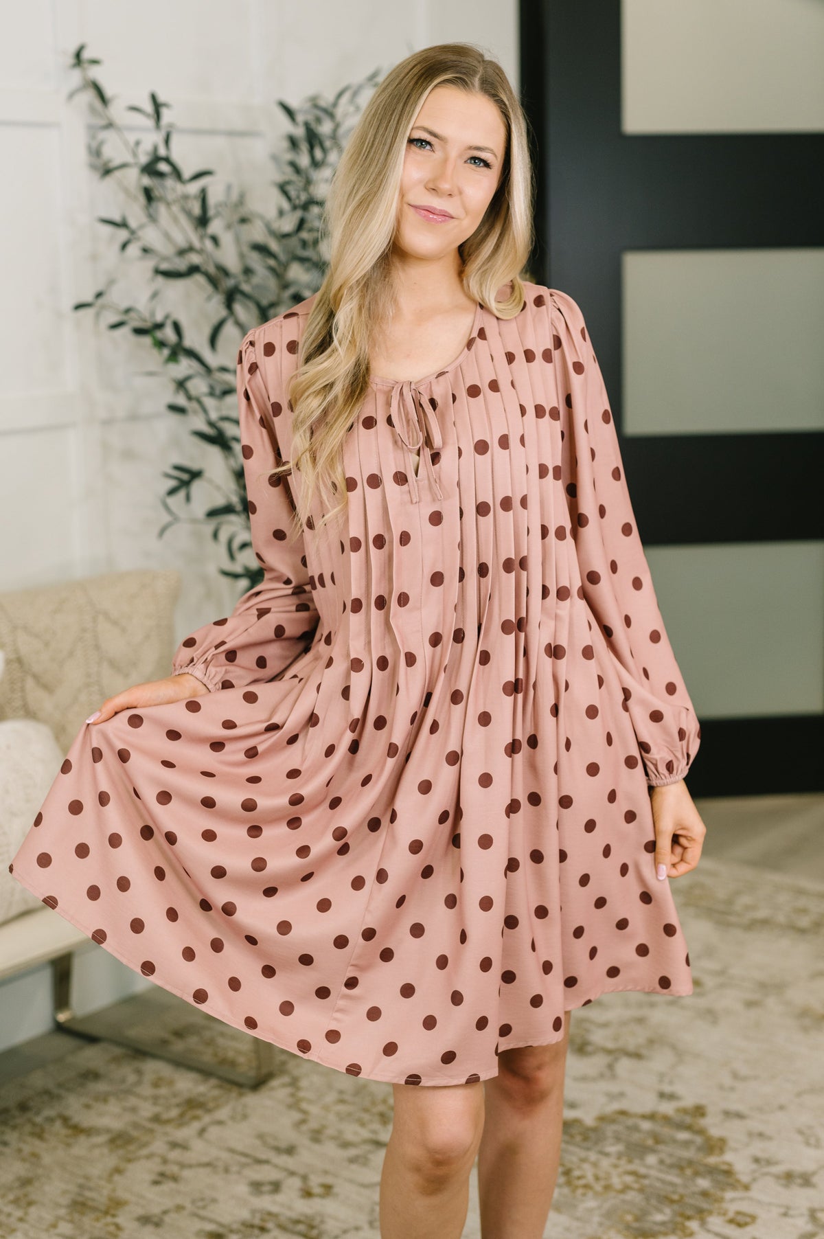 Woman wearing a pink polka dot dress with pleated details and a tie-neck, standing indoors.