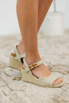 A close-up of a woman's foot wearing a beige linen wedge sandal with an adjustable buckle strap and a braided twine sole.