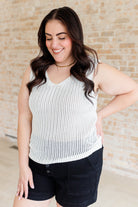 Woman wearing a white sleeveless top and black shorts against a beige brick wall.