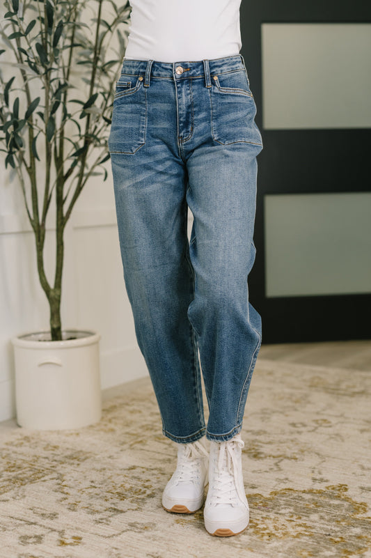 Woman wearing blue jeans and white sneakers standing in a room with a plant and striped wall.