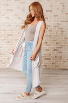 Woman wearing a beige top, light blue jeans, and a white cardigan against a brick wall.