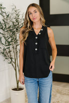 Woman wearing a black sleeveless top with buttons and blue jeans indoors.