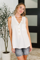 Woman wearing a white sleeveless blouse with buttons, standing indoors next to a plant.