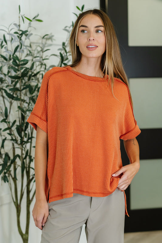 Woman wearing an orange ribbed sweater and beige pants standing indoors with a plant in the background.