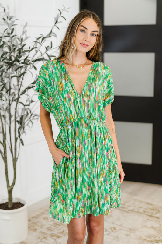 Woman wearing a green patterned dress standing indoors with a plant in the background