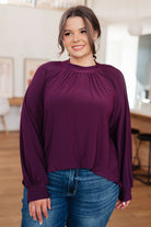 A model wearing a burgundy mock neck blouse with high-low hem and banded cuffs.