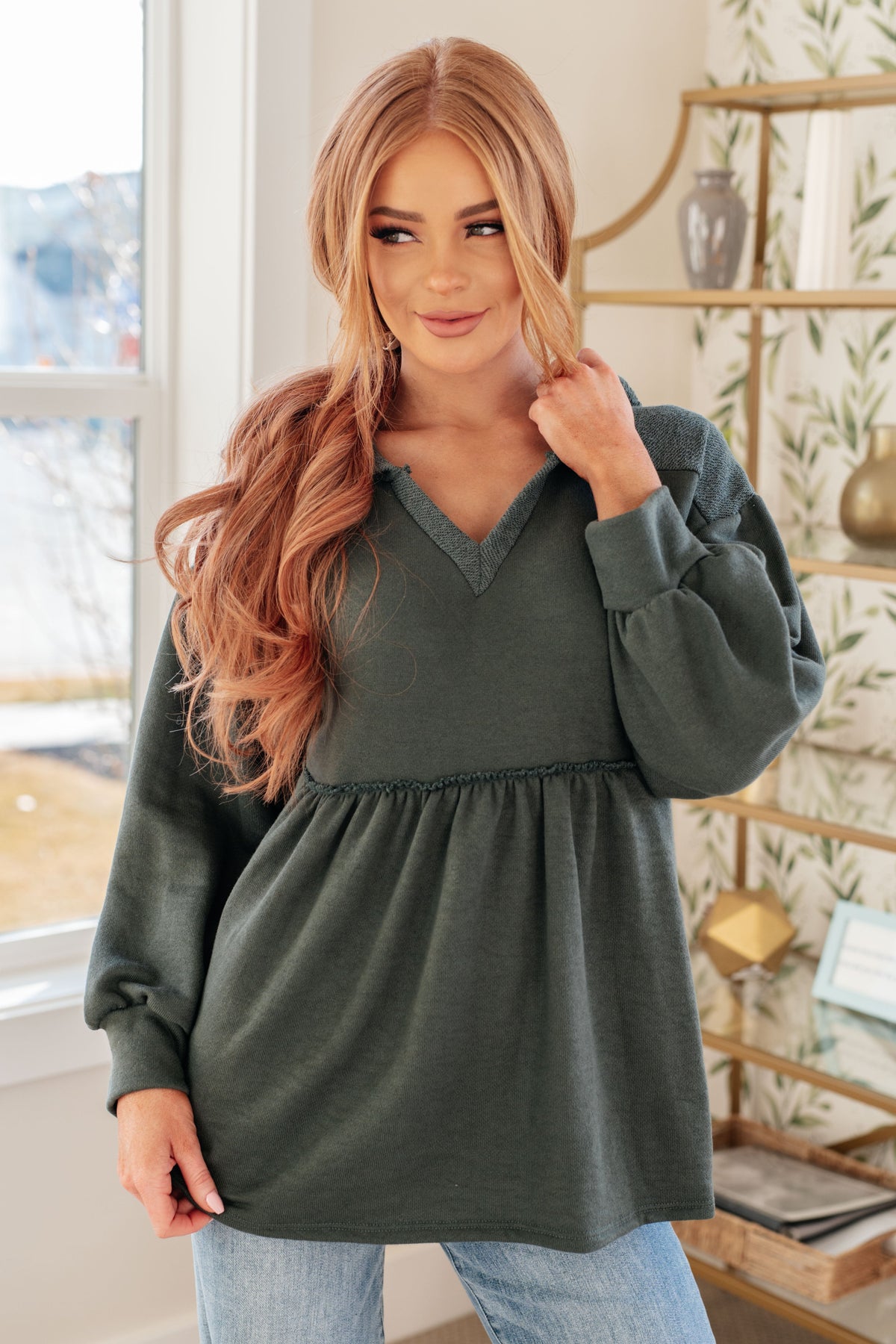 A woman wearing a forest green pullover with a hooded V-neck, gathered peplum, and balloon sleeves.