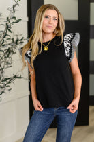 Woman wearing a black flutter sleeves top with floral  designs and blue jeans indoors.