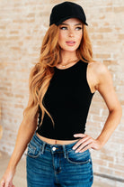 A woman wearing a black sleeveless rib knit tank top with a round neckline and cropped fit, paired with blue jeans and a black cap.