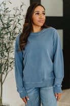 Woman wearing a blue sweatshirt and pants standing indoors with a plant in the background